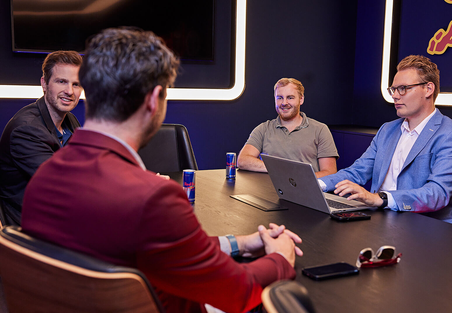 Vier mannen vergaderen in een moderne ruimte met sfeerverlichting, een laptop op tafel en een groot scherm op de achtergrond.